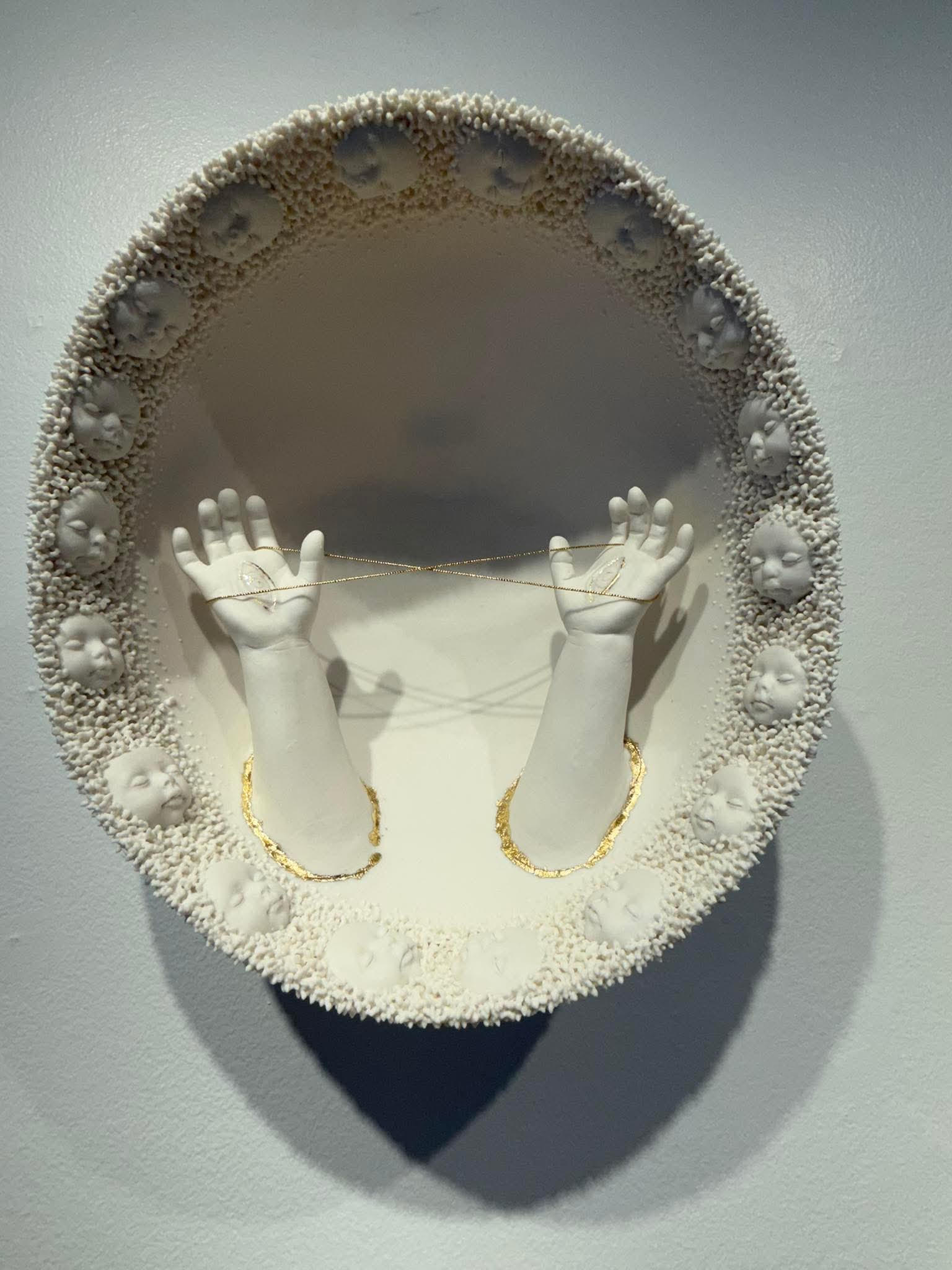 Oval porcelain wall sculpture with a textured border of child faces and two raised hands connected by a delicate gold chain